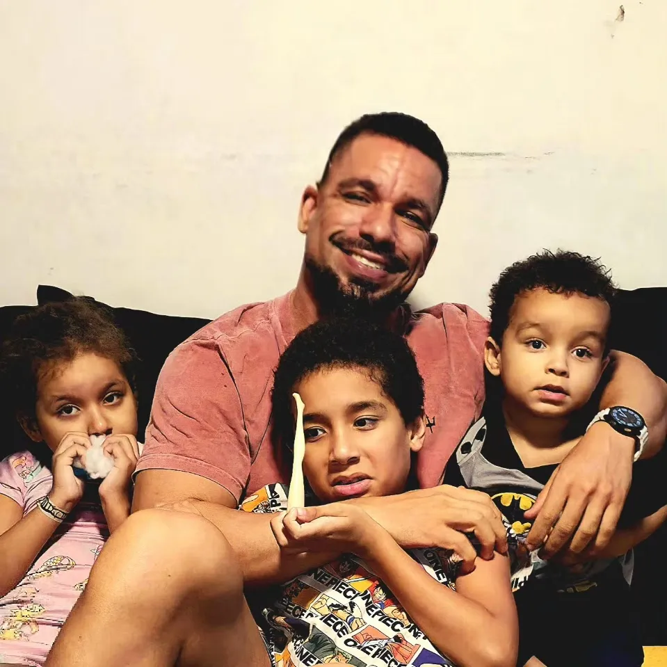 Rômulo Sutil sorrindo, sentado num sofá e abraçado a três crianças